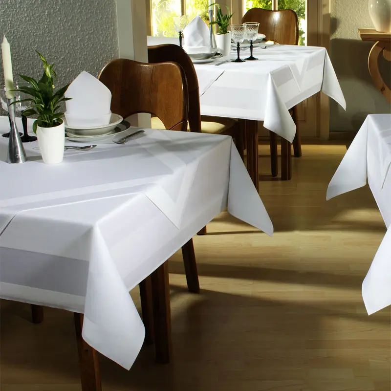 Das Bild zeigt einen elegant gedeckten Restaurant- oder Hotelbereich mit mehreren Tischen, die mit weißen Tischtüchern versehen sind. Die Tischwäsche hat eine dezente Atlaskante als Muster und wirkt hochwertig sowie klassisch. Auf den Tischen befinden sich weiße Stoffservietten, arrangiert zu einer spitzen Form, sowie Besteck, Teller, Gläser und kleine Dekorationselemente wie eine Pflanze und ein Kerzenständer. Die Stühle sind aus Holz und das Ambiente wird von Tageslicht aus dem Fenster erhellt, was eine freundliche und gepflegte Atmosphäre schafft.