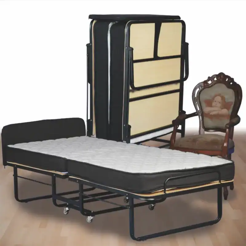Auf dem Bild ist ein Klappbett (auch Gästebett oder Zustellbett genannt) zu sehen. Im Vordergrund ist das Bett ausgeklappt: Es hat ein schwarzes Metallgestell mit Rollen, eine helle Holzoptik an den Seiten sowie eine dünne Matratze mit gestepptem Bezug. Dahinter steht das Bett im zusammengeklappten Zustand, wodurch es platzsparend verstaut werden kann. Rechts daneben befindet sich ein antiker Holzstuhl mit Armlehnen. Die Rückenlehne ist mit einem Polster bezogen, auf dem ein Engelmotiv abgebildet ist. Der Boden ist aus hellem Holzlaminat. Das Bild zeigt also ein praktisches Klappbett, das für Gäste genutzt werden kann und sich leicht verstauen lässt.