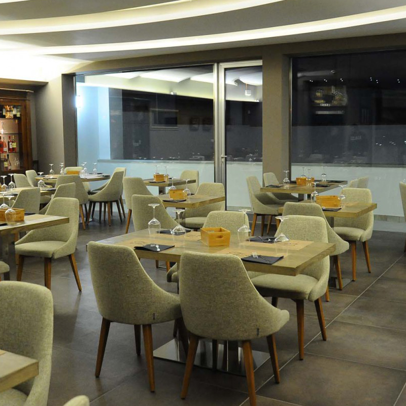 Armlehnstuhl Modell KONTEA 590 von Et al. – eleganter Polsterstuhl mit hoher Rückenlehne und Buchenholzgestell für moderne Restaurants und Hotel-Lounges - Hantermann - Der Hotelausstatter Mehrere KONTEA-590-Armlehnstühle in hellgrauem Stoff mit Holzbeinen in einem modernen Restaurant mit quadratischen Holztischen