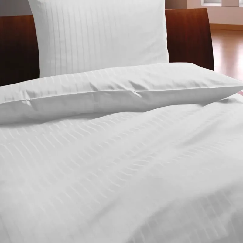 Das Bild zeigt ein elegant bezogenes Bett mit weißer Damast-Bettwäsche. Die Bettwäsche weist ein feines, vertikales Streifenmuster auf und wirkt besonders hochwertig und frisch. Der Bezug liegt glatt und ordentlich auf Bettdecke und Kissen und vermittelt einen professionellen, stilvollen Eindruck, wie er in Hotels oder gehobenen Gästezimmern üblich ist. Die helle Farbe und das dezente Design sorgen für ein modernes und einladendes Ambiente.