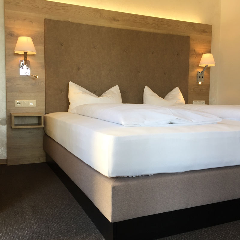 Hotel bedroom with a large padded headboard, two wall lamps, and a neatly made white bed.