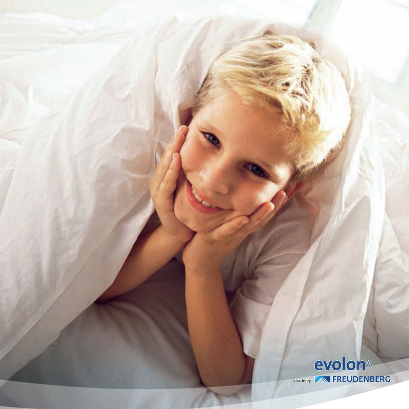 EVOLON® Allergikerbezug – optimaler Milbenschutz für Matratze, Decke und Kissen - Hantermann - Der Hotelausstatter Lächelndes Kind liegt unter einer weißen Bettdecke mit EVOLON Allergikerbezug, Symbol für milbendichten Schlafkomfort
