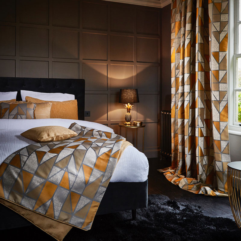 Geometric gold, gray, and taupe bedspread and matching curtains in a stylish hotel-style bedroom with a dark paneled wall and warm lighting.