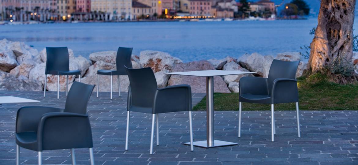 INOX 4421 Tischgestell by PEDRALI – Outdoor-Café-Setting am Wasser - Hantermann - Der Hotelausstatter Mehrere graue Armlehnstühle und quadratische Bistrotische mit INOX 4421 Tischgestell by PEDRALI auf einer Terrasse am Wasser vor mediterraner Häuserkulisse in der Dämmerung.