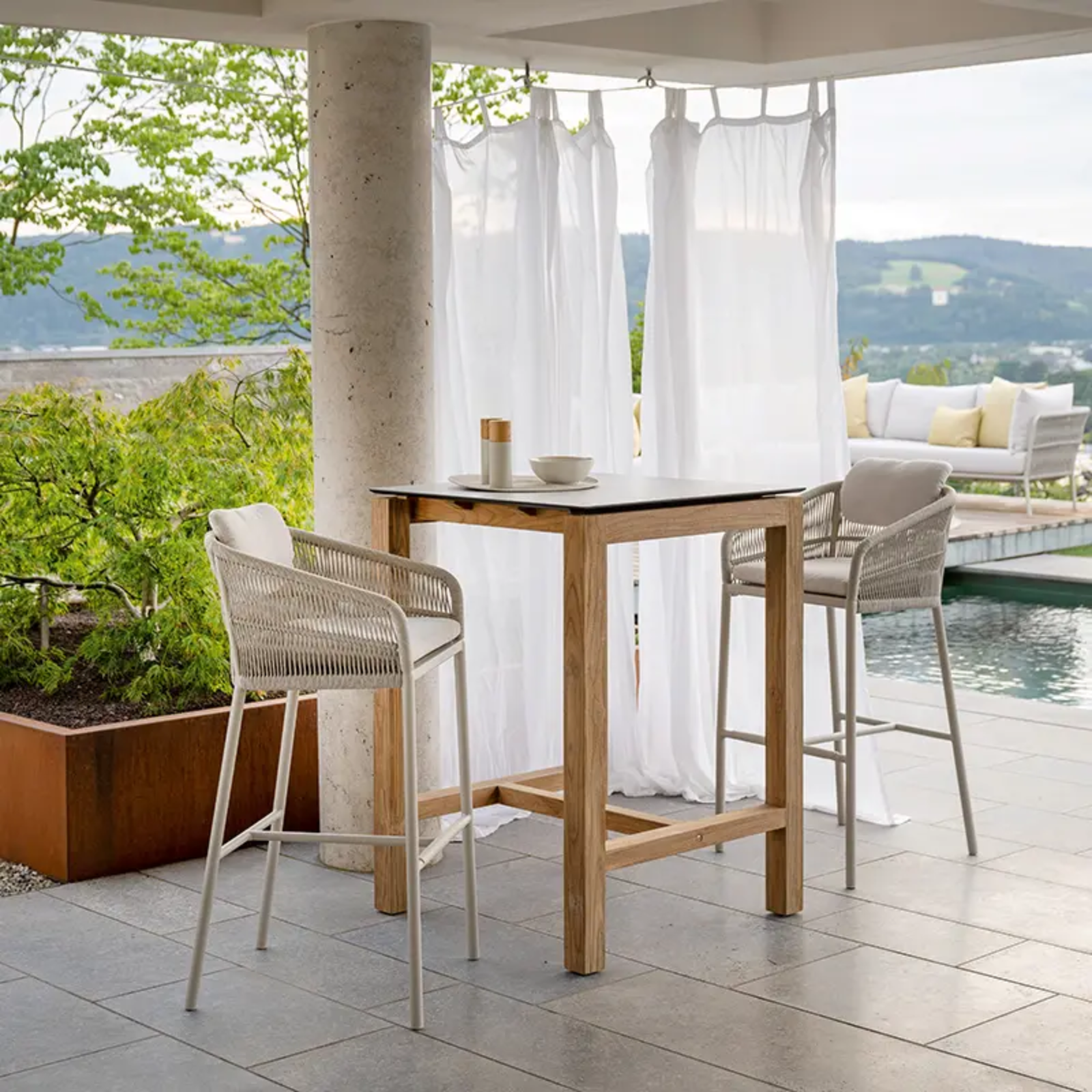 JELLA Barsessel greige/Kordel greige by STERN® – Outdoor-Barensemble mit Teak-Stehtisch - Hantermann - Der Hotelausstatter Zwei JELLA Barsessel greige/Kordel greige von STERN an hohem Teak-Stehtisch auf Terrasse mit Pool und Lounge-Sofas im Hintergrund