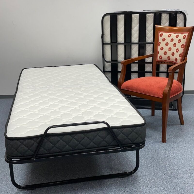 Klappbett (Gästebett) – Es steht aufgeklappt im Vordergrund. Das Bettgestell ist aus schwarzem Metall, und darauf liegt eine weiße, gesteppt wirkende Matratze mit einem dunkelgrauen Rand.