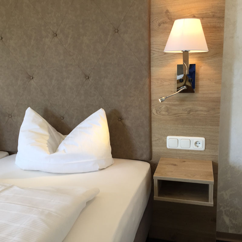 LYONBED Boxspringbett KÜMMERSBRUCK SUPERIOR – elegantes Hotelbett mit hohem Komfortkopfteil - Hantermann - Der Hotelausstatter Ausschnitt des LYONBED Boxspringbett KÜMMERSBRUCK SUPERIOR mit gesteppter Polsterkopfteilfläche, weißer Matratze und Wandlampe aus Holzpaneel