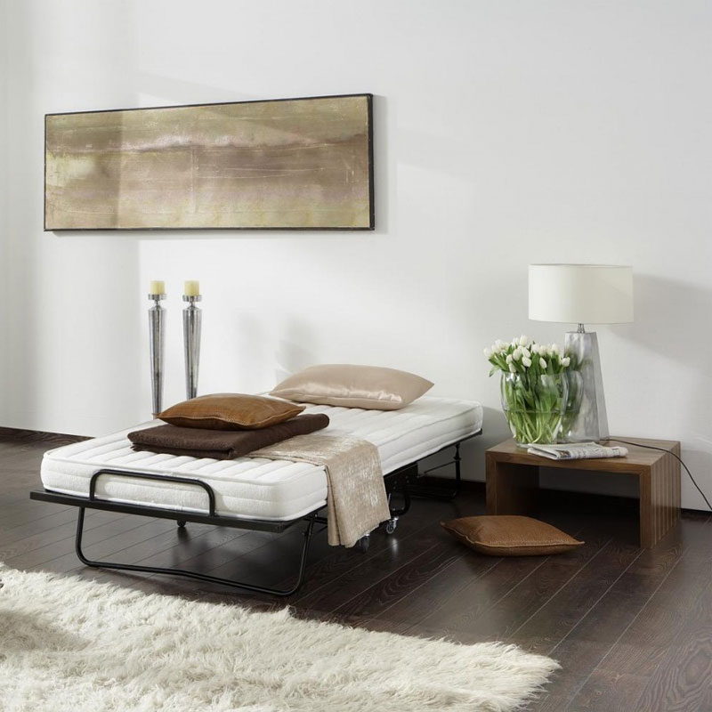 MANHATTAN Raumsparbett – aufgeklapptes Gästebett im modernen Zimmer - Hantermann - Der Hotelausstatter Aufgeklapptes MANHATTAN Raumsparbett mit weißer Matratze, Kissen und Plaid in hell eingerichtetem Zimmer mit Beistelltisch, Lampe und Wandbild