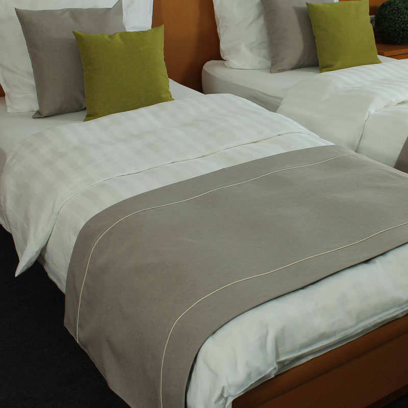 MIO Bettläufer/Fußplaid – dezentes Grau mit frischen grünen Dekokissen - Hantermann - Der Hotelausstatter Zwei Hotelbetten mit weißer Bettwäsche, grauem MIO Bettläufer und grauen sowie apfelgrünen Dekokissen