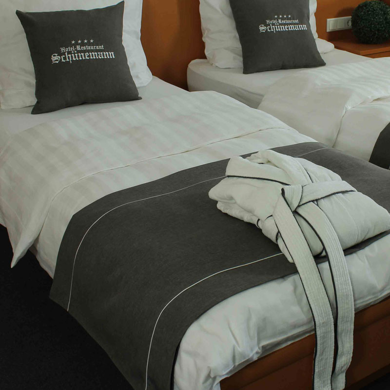 MIO Bettläufer/Fußplaid mit Logo-Kissen – stilvolle Präsentation von Hoteltextilien - Hantermann - Der Hotelausstatter Zwei Hotelbetten mit weißer Bettwäsche, anthrazitfarbenem MIO Bettläufer, Logo-Dekokissen und einem zusammengelegten Bademantel am Fußende