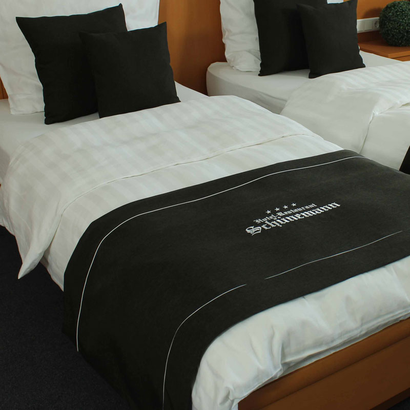 MIO Bettläufer/Fußplaid mit Logo – klassischer Schwarz-Weiß-Look für Hotel Schünemann - Hantermann - Der Hotelausstatter Zwei Hotelbetten mit weißer Bettwäsche, schwarzem MIO Bettläufer mit Hotel-Schünemann-Logo und passenden schwarzen Dekokissen