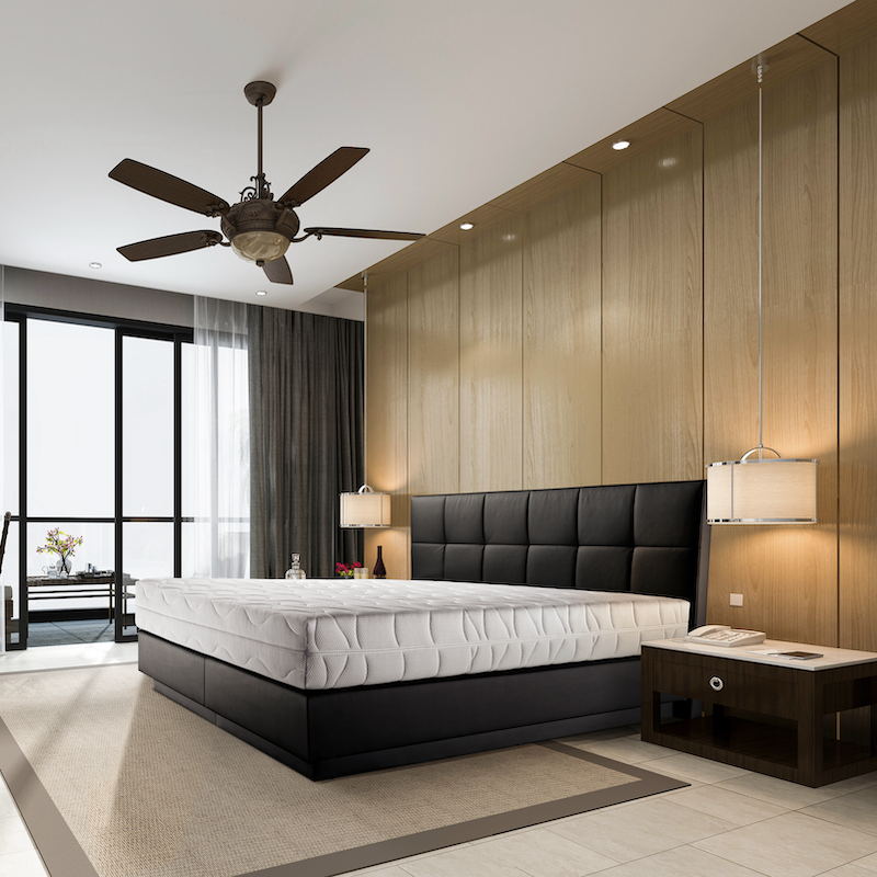 Modernes Hotelzimmer mit schwarzem Boxspringbett, hohem gepolsterten Kopfteil, Holzpaneelwand, Deckenventilator und Hängeleuchten