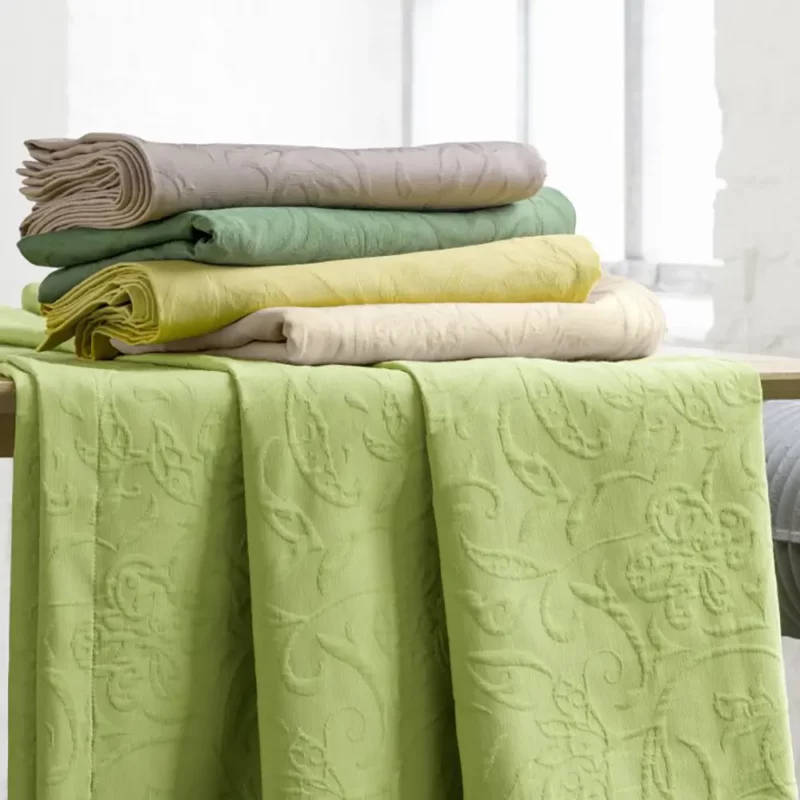 Das Bild zeigt mehrere farbige Tischdecken aus einer Baumwoll-Polyester-Mischung mit floraler, leicht erhabener Piqué-Struktur. Die Decken sind in hellen, natürlichen Farbtönen wie Grün, Gelb und Beige gehalten und dekorativ gefaltet auf einem Holztisch platziert. Im Vordergrund liegt eine grüne Tischdecke locker ausgebreitet, sodass die lebendige Struktur und das Muster gut sichtbar sind. Die Szenerie wirkt freundlich und modern und vermittelt einen hochwertigen, wohnlichen Eindruck.