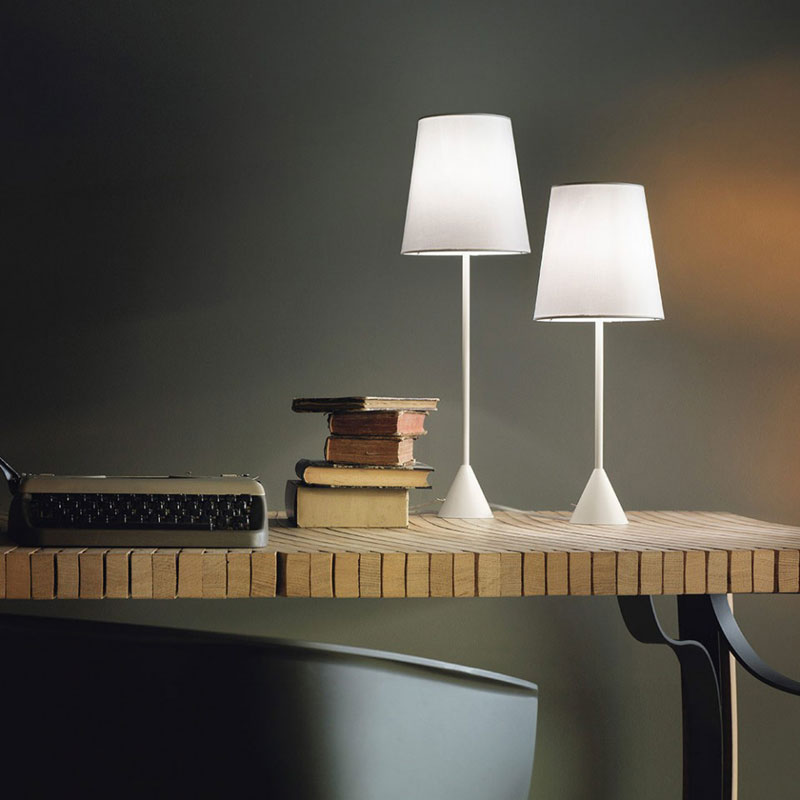 Nachttischlampe LYNDA – Moderne Tischleuchte in Weiß - Hantermann - Der Hotelausstatter Zwei moderne Nachttischlampen LYNDA in Weiß mit konischen Lampenschirmen, schlanken Leuchtenstäben und kegelförmigen Standfüßen auf einem Holztisch neben Büchern und einer Schreibmaschine