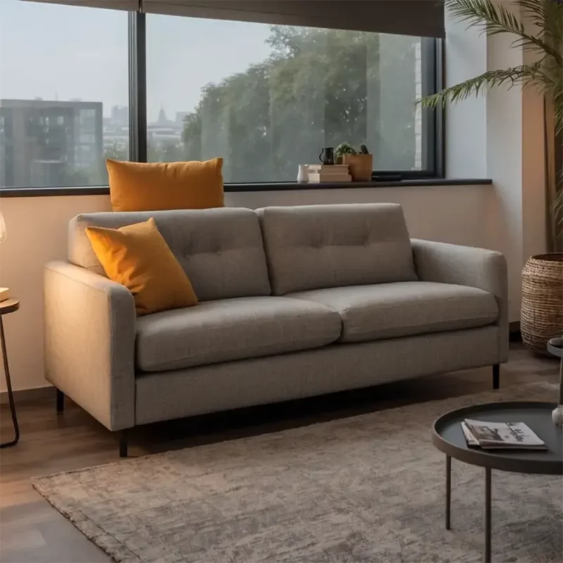 Auf dem Bild ist ein modernes, elegantes Schlafsofa in einem hellgrauen Stoffbezug zu sehen. Das Sofa steht in einem hellen, gemütlichen Wohnzimmer mit großen Fenstern, die viel Tageslicht hereinlassen. Zwei dekorative Kissen in einem warmen Gelbton setzen farbliche Akzente auf dem Sofa. Vor dem Sofa liegt ein dezenter Teppich in Grautönen, und daneben steht ein runder Couchtisch mit Zeitschriften darauf. Die Einrichtung wirkt schlicht und einladend, unterstrichen durch eine beleuchtete Lampe und einige Pflanzen, die für eine wohnliche Atmosphäre sorgen.