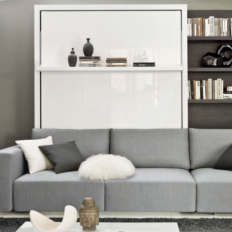 Schrankbett LEEDS im geschlossenen Zustand mit weißer Hochglanzfront, Wandboard mit Deko und grauem Sofa davor neben Bücherregal