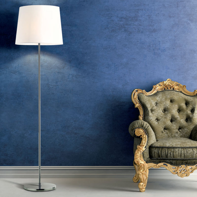 SOFT Bodenleuchte mit weißem, konisch geformtem Lampenschirm, schlankem silberfarbenem Metallgestell und rundem Standfuß neben einem klassischen Sessel vor blauer Wand