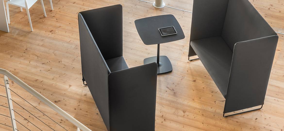 Stylus Tischgestell by PEDRALI – quadratischer Lounge- & Besprechungstisch - Hantermann - Der Hotelausstatter Quadratischer Tisch mit Stylus Tischgestell by PEDRALI, schwarzer Säule und breitem Rundfuß, arrangiert zwischen zwei hohen Lounge-Sitzmodulen in einem offenen Raum mit Holzboden.