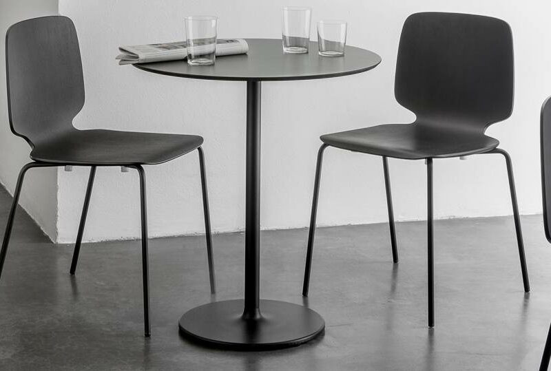 Runder Bistrotisch mit Stylus Tischgestell by PEDRALI, schwarzer Säule und rundem Fuß, umgeben von drei schwarzen Vierfußstühlen in einem minimalistischen Innenraum.