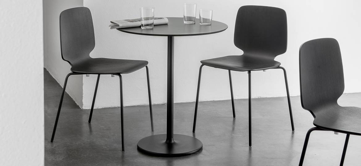 Stylus Tischgestell by PEDRALI – runder Bistrotisch im Minimal-Design - Hantermann - Der Hotelausstatter Runder Bistrotisch mit Stylus Tischgestell by PEDRALI, schwarzer Säule und rundem Fuß, umgeben von drei schwarzen Vierfußstühlen in einem minimalistischen Innenraum.