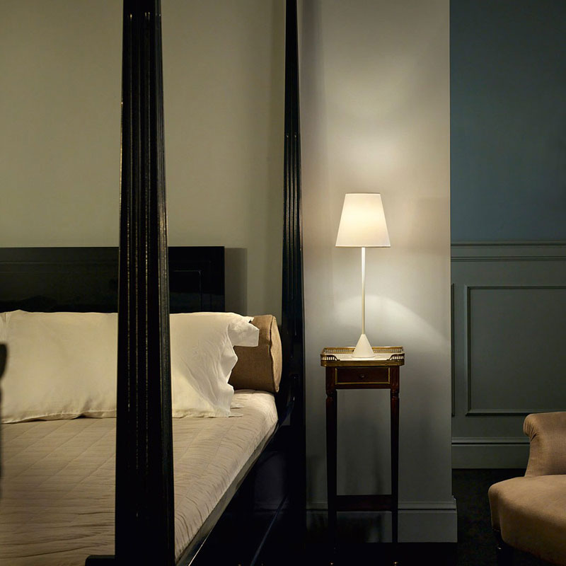 Weiße Nachttischlampe LYNDA mit konischem Lampenschirm und schlankem Leuchtenstab auf einem eleganten Nachttisch neben einem Hotelbett mit weißen Kissen