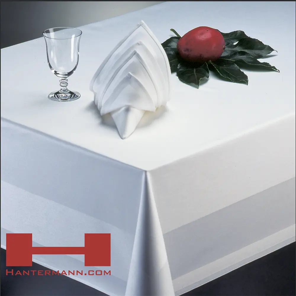 Das Bild zeigt einen elegant gedeckten Tisch mit einem weißen Tischtuch, darauf eine kunstvoll gefaltete Stoffserviette in weiß, ein sowie ein kleines Glas mit Stiel. Rechts daneben befindet sich ein roter Apfel auf einem grünen Blatt. In der unteren linken Ecke ist das Logo und der Schriftzug "HANTERMANN.COM" in Rot abgebildet. Das gesamte Arrangement vermittelt einen hochwertigen und stilvollen Eindruck, passend für gehobene Gastronomie oder Hotellerie.