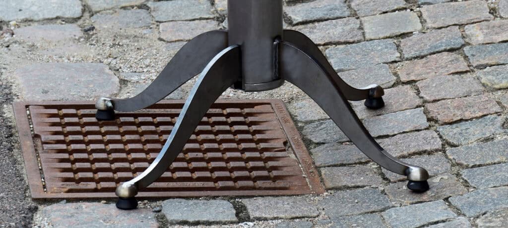 Das Bild zeigt den unteren Teil eines StableTable Metalltischgestells mit vier geschwungenen Beinen, die jeweils mit runden schwarzen Gummifüßen versehen sind. Der Tisch steht im Freien auf einem Pflastersteinboden. Unter einem der Tischbeine befindet sich ein quadratischer, gitterartiger Metalldeckel, vermutlich ein Abfluss- oder Versorgungsschacht. In der Mitte des Bildes steht in großer, weißer Schrift das Wort: „Innovationen“ Die Ecken des Bildes sind leicht abgerundet, was ihm einen gestalterischen Rahmen verleiht.