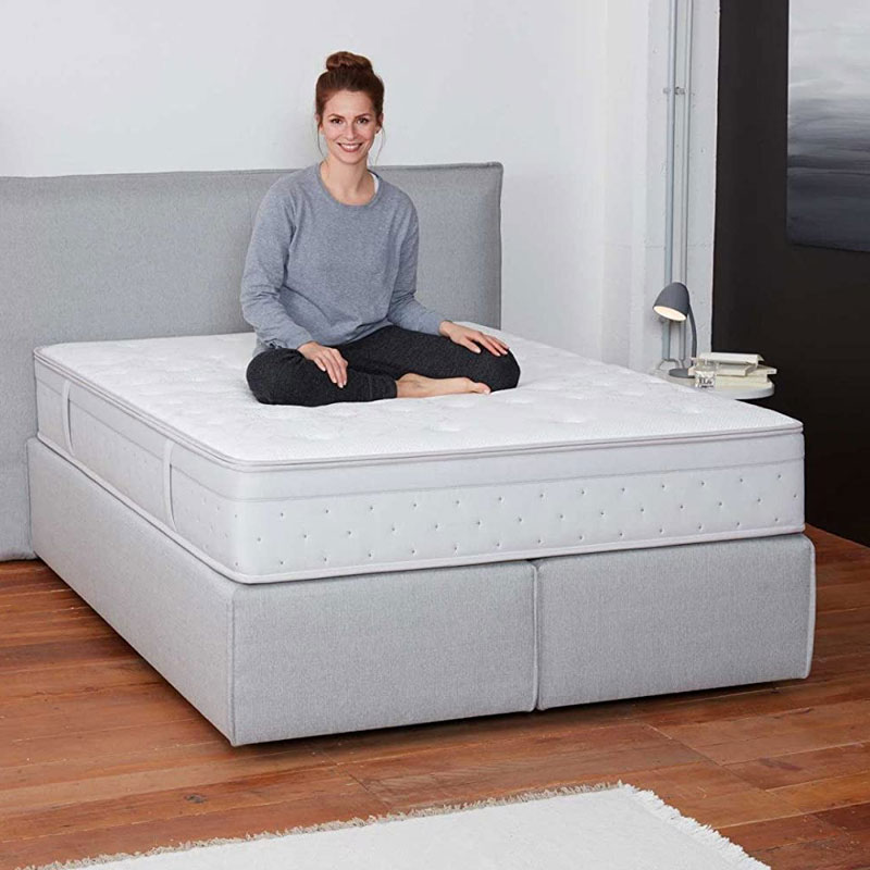 Frau sitzt im Schneidersitz auf einem hellgrauen Boxspringbett mit hoher, weißer Matratze in modernem Schlafzimmer mit Holzfußboden und Nachttisch mit Lampe.