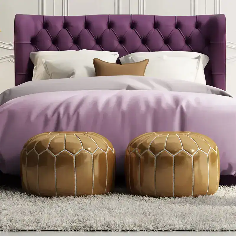 Ein Box spring Bett in der Größe 180 × 200 cm mit einem gehefteten geschwungenen Kopfteil in der Farbe Lila. Auf dem Box spring Bett liegen auf jeder Seite 2 Kissen und ein kleines sowie eine Bettdecke und vor dem Bett befinden sich zwei runde Lederhocker.