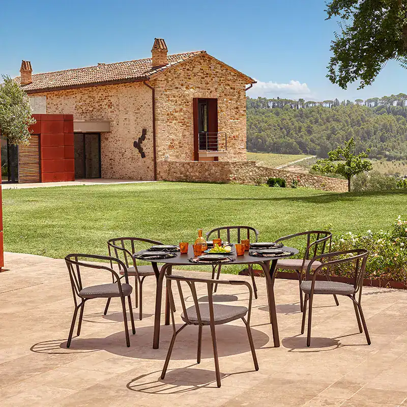 Terrassenstühle stehen um einen Tisch auf einer Steinfläche vor einem grünen Rasen mit toskanischem Steinhaus und blauem Himmel im Hintergrund.