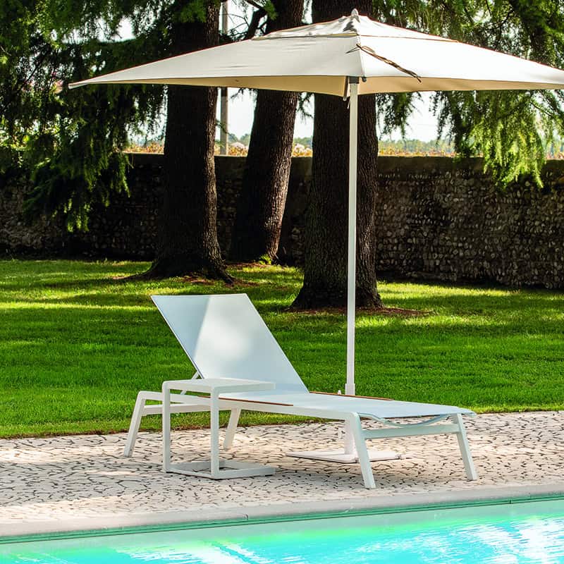 weiße Sonnenliege am Pool vor grünem rasen unter Sonnenschirm, dahinter Bäume.