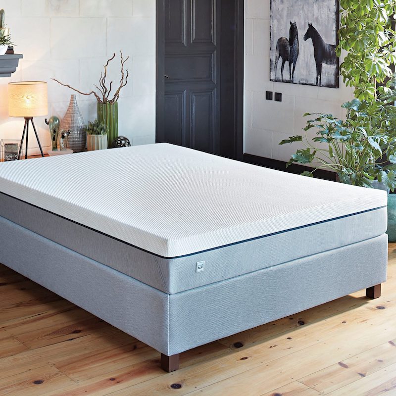 Das Bild zeigt ein modernes Boxspringbett in einem hellen, stilvoll eingerichteten Schlafzimmer. Das Bett hat einen hellgrauen Unterbau mit Holzfüßen und eine weiße, glatte Matratzenoberfläche mit feiner Struktur. Im Hintergrund stehen auf einem Sideboard eine Lampe mit warmem Licht, dekorative Vasen und Pflanzen, was dem Raum eine gemütliche Atmosphäre verleiht. An der Wand hängt ein Bild mit zwei Pferden, daneben befindet sich eine dunkle Tür; rechts vom Bett stehen hohe, üppige Grünpflanzen auf einem Holzfußboden.
