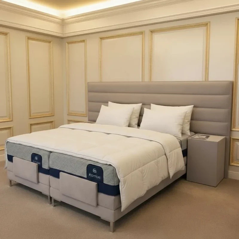Das Bild zeigt ein repräsentatives, luxuriös eingerichtetes Schlafzimmer mit einem hochwertigen Boxspringbett aus der Casaletto Luxury Suite Collection. Das Bett besitzt ein großes, gepolstertes Kopfteil in dezentem Grau und ist sorgfältig mit einem weißen Bettlaken, zwei separaten Daunendecken sowie jeweils zwei Kissen pro Seite bezogen. Links und rechts vom Bett steht jeweils ein moderner, schlichter Nachtisch mit einer Zeitschrift darauf. Die Wandgestaltung setzt auf helle Flächen mit goldfarbenen, klassisch inspirierten Zierleisten, die dem Raum Eleganz und Hotelambiente der Premium-Kategorie verleihen. Der Teppichboden und die indirekte Beleuchtung unterstreichen die behagliche und exklusive Atmosphäre auf höchstem Niveau.