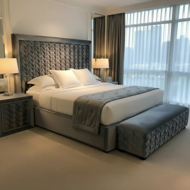 Das Bild zeigt das elegante Boxspringbett Nova aus der Luxury Suite Collection, präsentiert von Hantermann - Der Hotelausstatter aus Kleinostheim. Das Bett besticht durch sein markantes Kopfteil, dessen grauer Stoff kunstvoll mit einem Blattmuster strukturiert und sorgfältig gepolstert ist. Die Bettbank am Fußende greift das Dekor und das Material des Kopfteils wieder auf und sorgt für eine harmonische, durchdachte Raumgestaltung. Flankiert wird das Bett von zwei stilvollen Nachttischen mit passenden Lampen, die das schlichte und edle Ambiente zusätzlich unterstreichen. Der Raum ist lichtdurchflutet durch bodentiefe Fenster, deren Vorhänge dem modernen Luxuslook entsprechen. Das Farbkonzept ist klar: Grautöne, Weiß und dezente Beige-Nuancen schaffen eine entspannte und exklusive Schlafumgebung. Die hochwertigen Materialien und das zeitlose Design machen dieses Bett ideal für gehobene Hotelsuiten und anspruchsvolle Gäste.