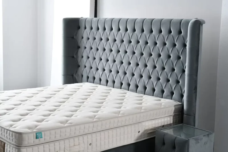 Das Bild zeigt einen luxuriösen Ausschnitt eines Boxspringbettes, das den Namen „Grand_Palais_Royal“ trägt. Im Fokus steht das beeindruckende, hohe Kopfteil mit großzügiger Chesterfield-Steppung in elegante, samtartige Stoff-Optik und dezentem Grauton. Die gewählte Polsterung wirkt massiv, weich und vermittelt königlichen Komfort. Die Matratze ist ungewöhnlich hoch und wirkt besonders luxuriös – ihre Kanten sind präzise abgesteppt, mit einem Gittermuster in Cremeweiß und filigraner Einfassung. Die sichtbaren Haltegriffe und Belüftungsösen deuten auf hochwertige Verarbeitung hin. rechts neben dem Bett befindet sich ein Nachttisch, ebenfalls in derselben gepolsterten Stoffoptik, mit einer Glasplatte als Oberfläche – ein Design, das perfekt auf das Gesamtbild abgestimmt ist. Der Hintergrund ist hell gehalten, die natürliche Lichtführung wirkt wohnlich und betont die Exklusivität dieses Schlafmöbels.