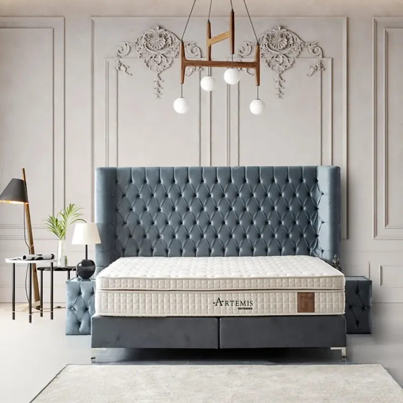 Das Bild zeigt ein luxuriös eingerichtetes Schlafzimmer, das durch zeitloses Design und hochwertige Materialien besticht. Im Mittelpunkt steht ein imposantes Bett mit einem sehr hohen, dunkelgrau gepolsterten Kopfteil in eleganter Chesterfield-Steppung. Das Bettgestell ist vollständig mit einem opulenten, hochformatigen Matratzenmodell in heller, cremefarbener Optik abgedeckt, wobei das Label "Artemis" auf der Matratze auf eine exklusive Qualitätsmarke hinweist. Hintergrund und Raumdesign Die Wände sind mit aufwendigen, klassizistischen Zierleisten und floralen Ornamenten versehen, was dem Raum eine edle, fast palastartige Atmosphäre verleiht. Die Farbgebung der Wände bewegt sich im Spektrum heller Beige- und Grautöne und harmoniert perfekt mit dem sandfarbenen, hochwertigen Teppichboden, der den gesamten Raum bedeckt. Dadurch entsteht ein stimmiges und warmes Gesamtambiente. Möblierung und Details Rechts und links vom Bett befinden sich gepolsterte, kubische Nachttische, die das Designkonzept des Kopfteils aufnehmen. Auf der linken Bildseite steht eine kleine, elegante Leseecke mit einer modernen Stehlampe, einem weißen Beistelltisch und einer Zimmerpflanze, was für zusätzliche Wohnlichkeit sorgt. Die minimalistische Leuchte an der Decke, bestehend aus Holz und weißen Kugellichtern, rundet das stilvolle Gesamtbild ab und setzt einen dezenten, modernen Akzent. Insgesamt vermittelt das Zimmer Exklusivität, Komfort und zeitlose Eleganz – ideal für gehobene private Schlafräume oder exklusive Hotelsuiten.