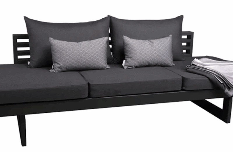 Das Bild zeigt eine moderne Outdoor-Lounge oder Liege mit einem klaren, minimalistischen Design. Beschreibung der Möbel Die Liege besteht aus einem stabilen, dunkelgrauen Metallgestell mit drei komfortablen Sitzpolstern in einem passenden Anthrazitton. Die Rückenlehne zeigt ein schlichtes Lattenmuster, das einen zeitgemäßen Look unterstreicht. Für zusätzlichen Komfort sorgen zwei große Rückenkissen sowie zwei kleinere rechteckige Zierkissen in einem helleren Grau mit feinem Muster. Rechts auf der Liege liegt eine gefaltete Decke oder ein Plaid in Grau und Weiß, was dem Ensemble ein wohnliches, einladendes Flair verleiht. Stil und Einsatzbereich Die Gesamtkomposition wirkt modern und hochwertig, und die Liege eignet sich besonders für den Einsatz im Außenbereich, etwa auf einer Terrasse, im Garten oder auf einem Balkon. Das schlichte, geradlinige Design macht sie zum idealen Möbelstück für stilvolle Hotelanlagen, moderne Wohnumgebungen oder luxuriöse Outdoor-Lounges.