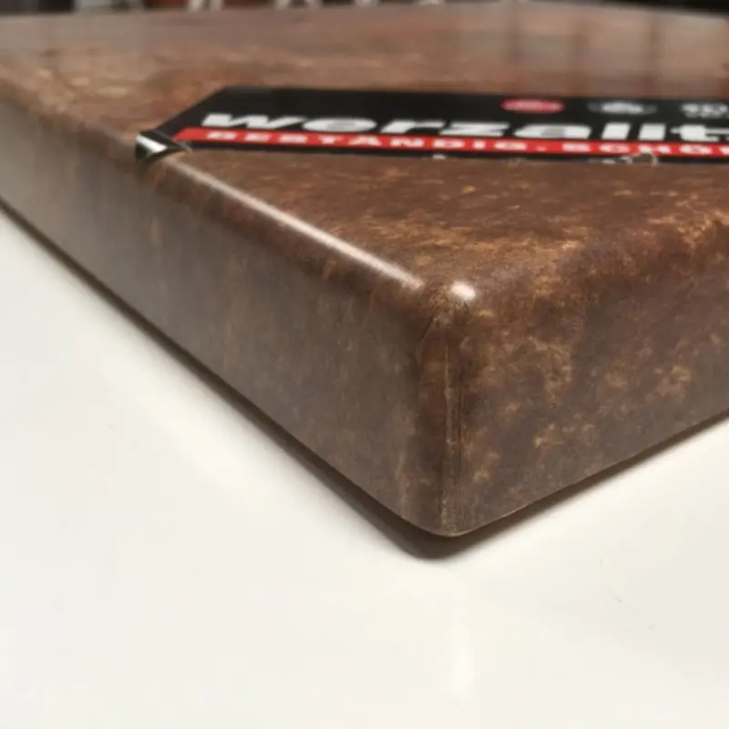 Das Bild zeigt eine Detailaufnahme der Ecke einer Werzalit-Tischplatte mit der Dekorvariante „Vulcano“. Die Oberfläche ist in einem marmorierten, erdigen Braun gehalten, wirkt robust und pflegeleicht. Die Kante ist sanft abgerundet und zeigt die charakteristische Verschmelzung von Dekorschicht und Kernmaterial, was für Stabilität und Widerstandsfähigkeit sorgt. Im oberen Bereich ist ein Ausschnitt eines Werzalit-Etiketts mit Hinweisen auf die Beständigkeit und Qualität des Produktes zu sehen.