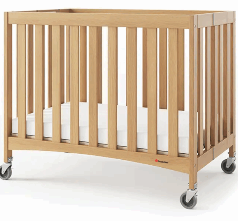 Das Bild zeigt ein kompaktes Babybett aus Holz mit einem klaren, minimalistischen Design. Material & Farbe: Natürliches Holz mit glatter Oberfläche und warmem Finish – wirkt hochwertig und freundlich Gitterstruktur: Alle vier Seiten sind mit vertikalen Holzstreben versehen, die für Sicherheit und gute Luftzirkulation sorgen Mobilität: Jedes Bein ist mit einem stabilen Rollenrad ausgestattet – insgesamt vier Rollen, zwei davon vermutlich feststellbar Matratze: Eine weiße Matratze liegt passgenau im Bett, wirkt sauber und bequem Das Bett ist für Kleinkinder gedacht, ideal für Hotels, Kitas oder den mobilen Einsatz. Die Kombination aus Funktionalität und schlichtem Design macht es besonders attraktiv für professionelle Umgebungen.