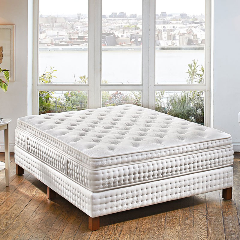 Helles Boxspringbett mit hoher, weißer, aufwendig gesteppten Matratze in einem Zimmer mit Holzboden vor bodentiefen Fenstern mit Blick über die Dächer einer Stadt.