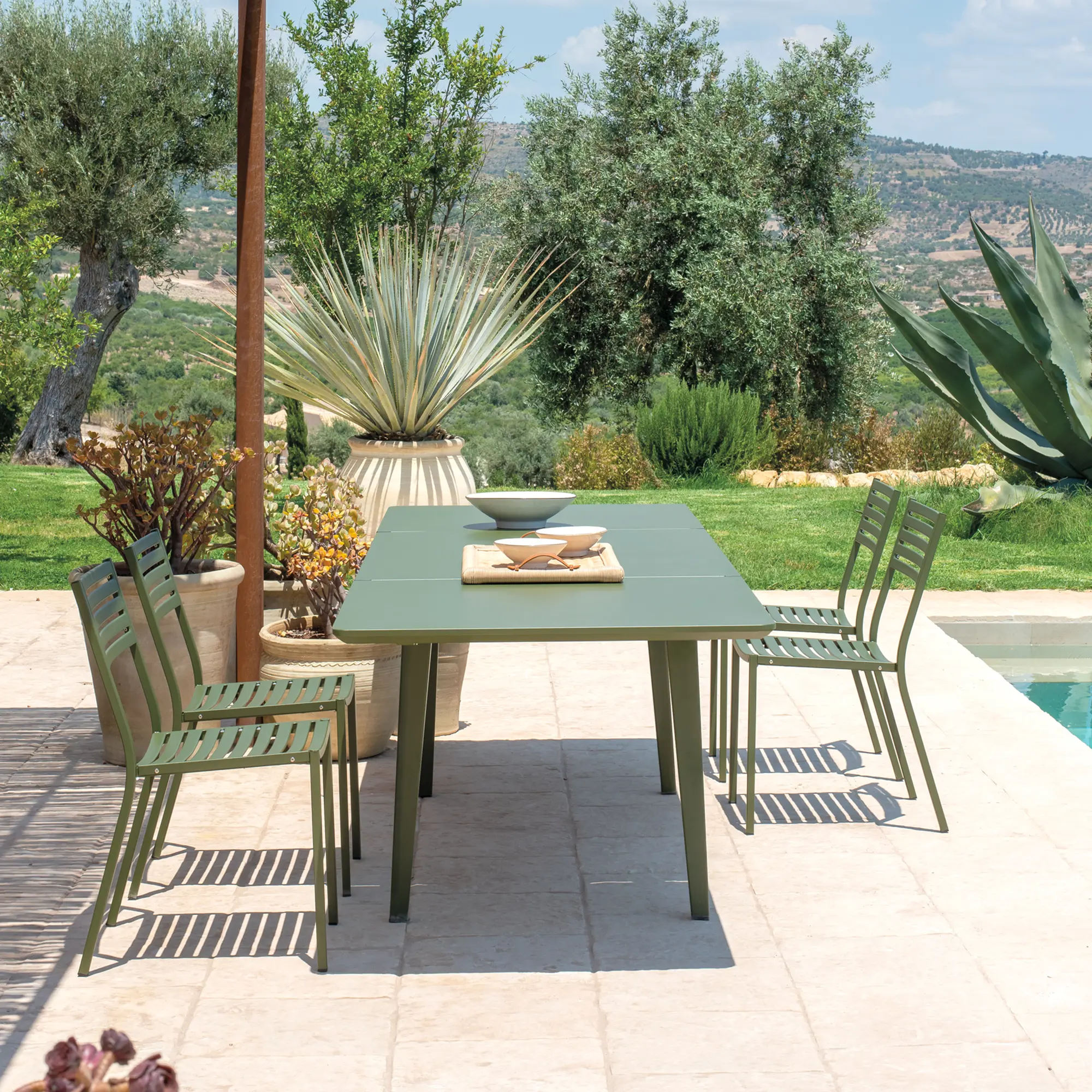 EMU SEGNO 263 Stapelstühle auf mediterraner Terrasse - Hantermann - Der Hotelausstatter Mediterrane Terrasse mit grünem Outdoor-Esstisch und vier EMU SEGNO 263 Stapelstühlen in Farbe Grün (17) aus Stahl mit Latten-Sitz und -Rückenlehne, platziert am Pool mit Blick in die Landschaft.