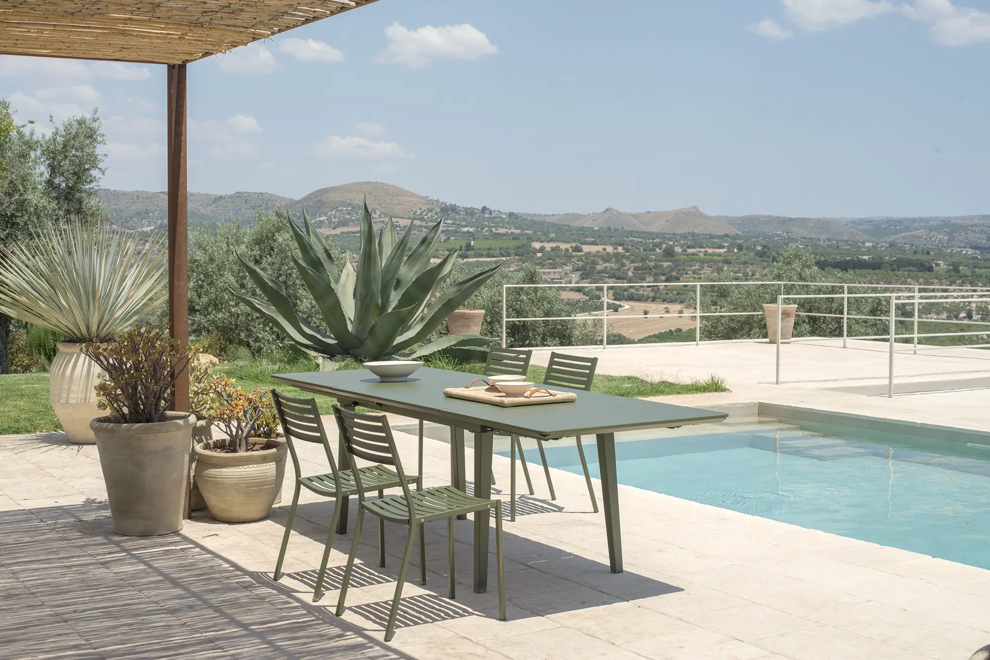 EMU SEGNO 263 Stapelstühle am mediterranen Pool - Hantermann - Der Hotelausstatter Modernes Pool-Ambiente mit grünem Outdoor-Esstisch und vier EMU SEGNO 263 Stapelstühlen aus Stahl, mediterrane Terrasse mit Landschaftsblick und großen Kübelpflanzen.