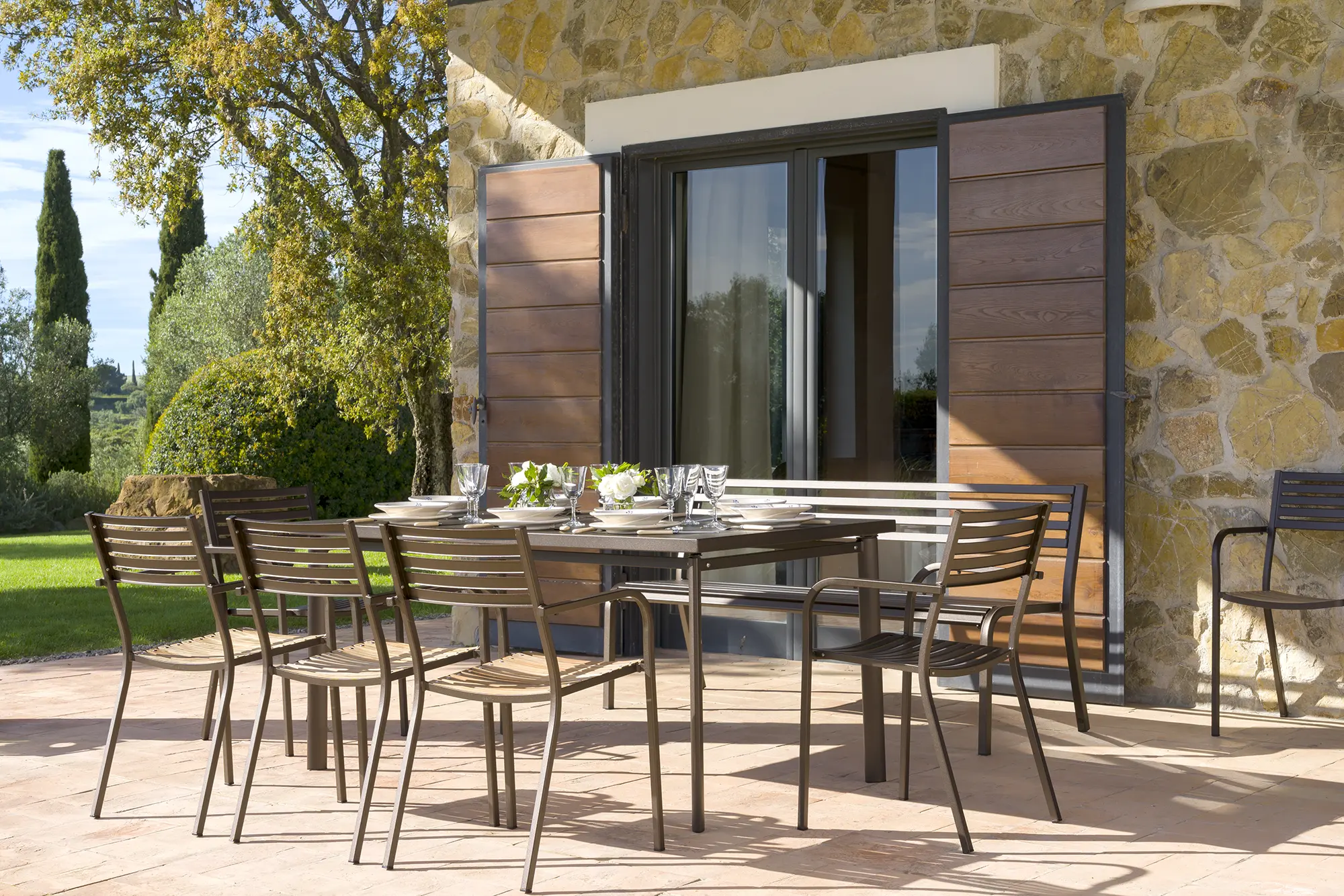 EMU-Segno-268-Stapelsessel-Ambiente-01 - Hantermann - Der Hotelausstatter Terrassen-Ambiente mit gedecktem Esstisch und mehreren EMU SEGNO 268 Stapelsesseln aus Stahl, modernes Outdoor-Mobiliar vor Natursteinfassade mit Gartenblick.