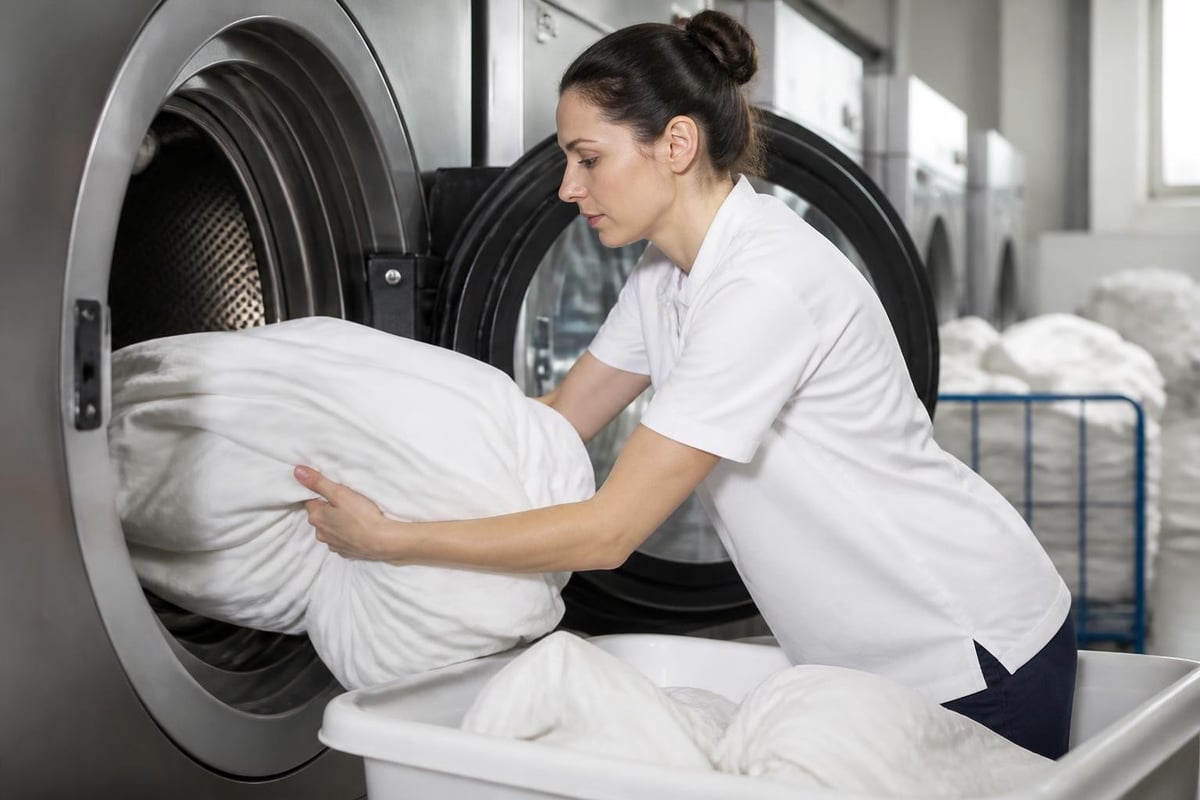 Mitarbeiterin in einer Wäscherei belädt eine große Waschmaschine mit weißen Bettwaren